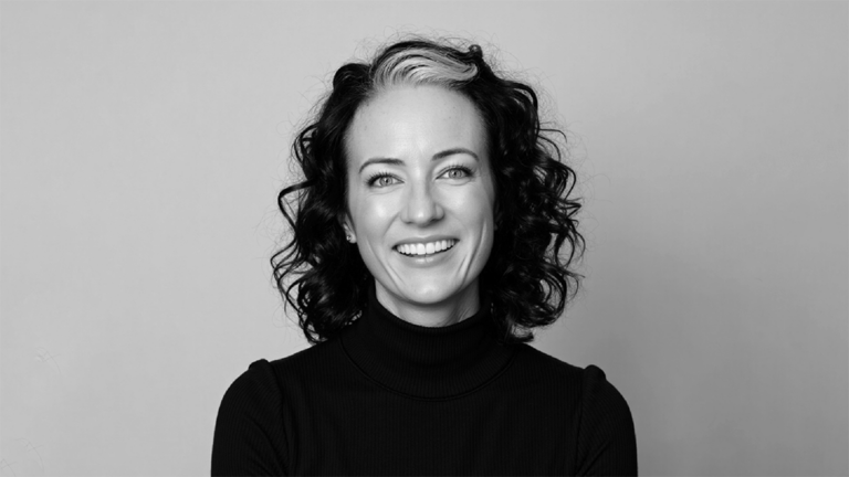 Head-and-shoulders portrait of a smiling woman with dark, curly hair and a light streak, wearing a black turtleneck against a neutral gray background.