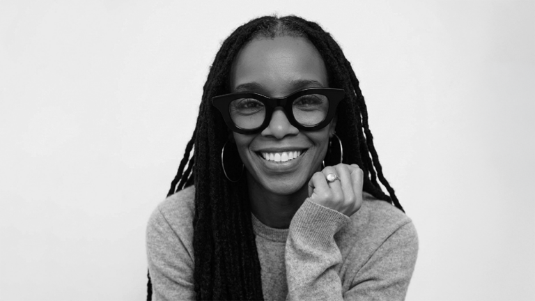 Portrait of a smiling Black woman with long dreadlocks, oversized black glasses, hoop earrings, and a gray sweater.