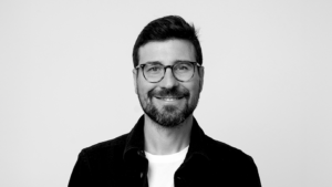 Smiling man with short dark hair and glasses, wearing a black jacket over a white T-shirt, facing the camera (black-and-white photo).