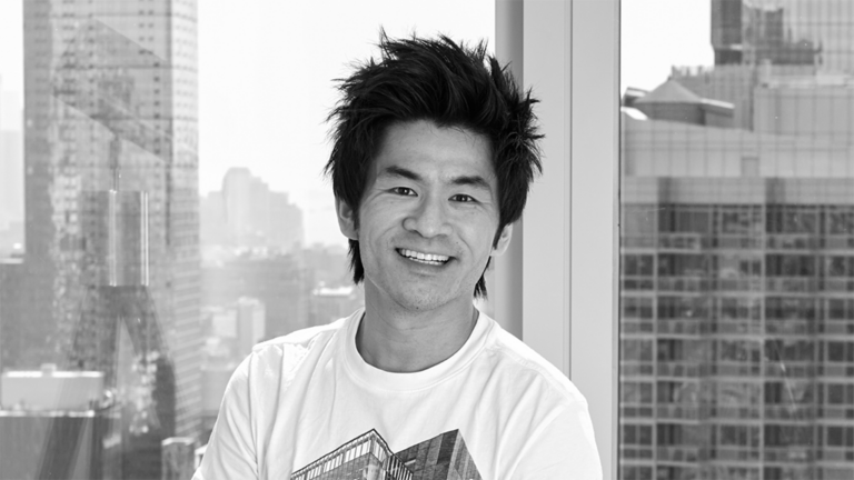 Smiling Asian man standing by a window in a high-rise office with a city skyline behind him (black and white photo).