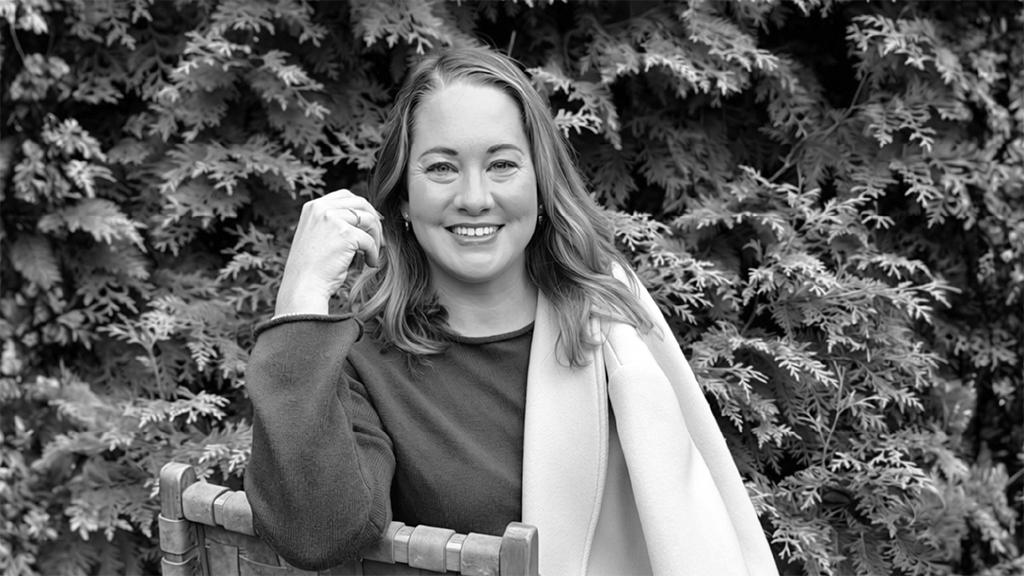 Smiling woman seated on a wooden bench with dense green foliage behind her in an outdoor setting (black and white). Clean, friendly portrait.
